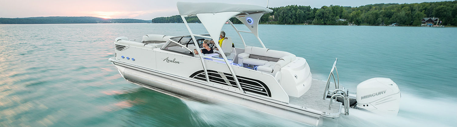 Two people on a 2018 Avalon Pontoons Excalibur dashing through a lake surrounded by trees.